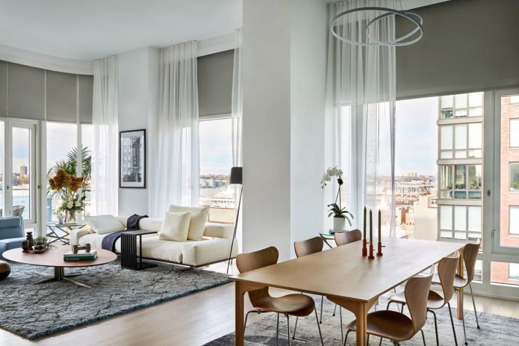 This luxurious Manhattan penthouse boasts floor-to-ceiling windows on every wall, adorned with beige blinds and sheer white curtains. The living area is nestled in one corner, featuring a white sofa with matching cushions and a stylish dark blue throw. A light blue armchair sits alongside the sofa, creating a cosy seating arrangement. An oval light wood coffee table showcases books and ornaments, resting on a plush grey/blue silk fluffy rug. In the corner, a vibrant bunch of flowers and tropical leaves complement the framed art on the wall. Adjacent to the living space, the dining area showcases a light wood dining table and chairs, adorned with candles, all placed on a blue/grey silk rug. A stunning white chandelier hangs above the table, while a white orchid graces a side table nearby.