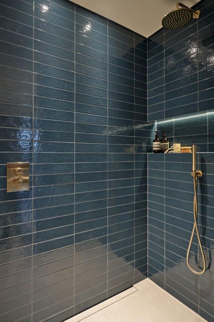 up-close image of a shower. The shower showcases deep blue subway tiles from the prestigious Collection Refoli by Marca Corona, exuding elegance and style. The gold brushed brass hardware, including the shower head and control panel, adds a touch of sophistication. Complementing the shower, light cream floor tiles create a striking contrast. A built in shelf halfway up the wall with built in lights, illuminating the shelf and displaying an array of shower products.
