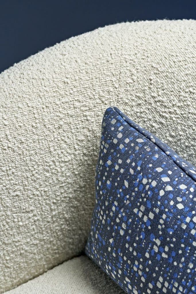 Indulge in the luxury of a zoomed-in image showcasing an armchair adorned with exquisite cream boucle fabric from CB2 America. The attention to detail is evident in the accent cushion, crafted from a dark blue fabric adorned with intricate patterns of light blue and beige. The combination of textures and colours creates a captivating and sophisticated aesthetic..