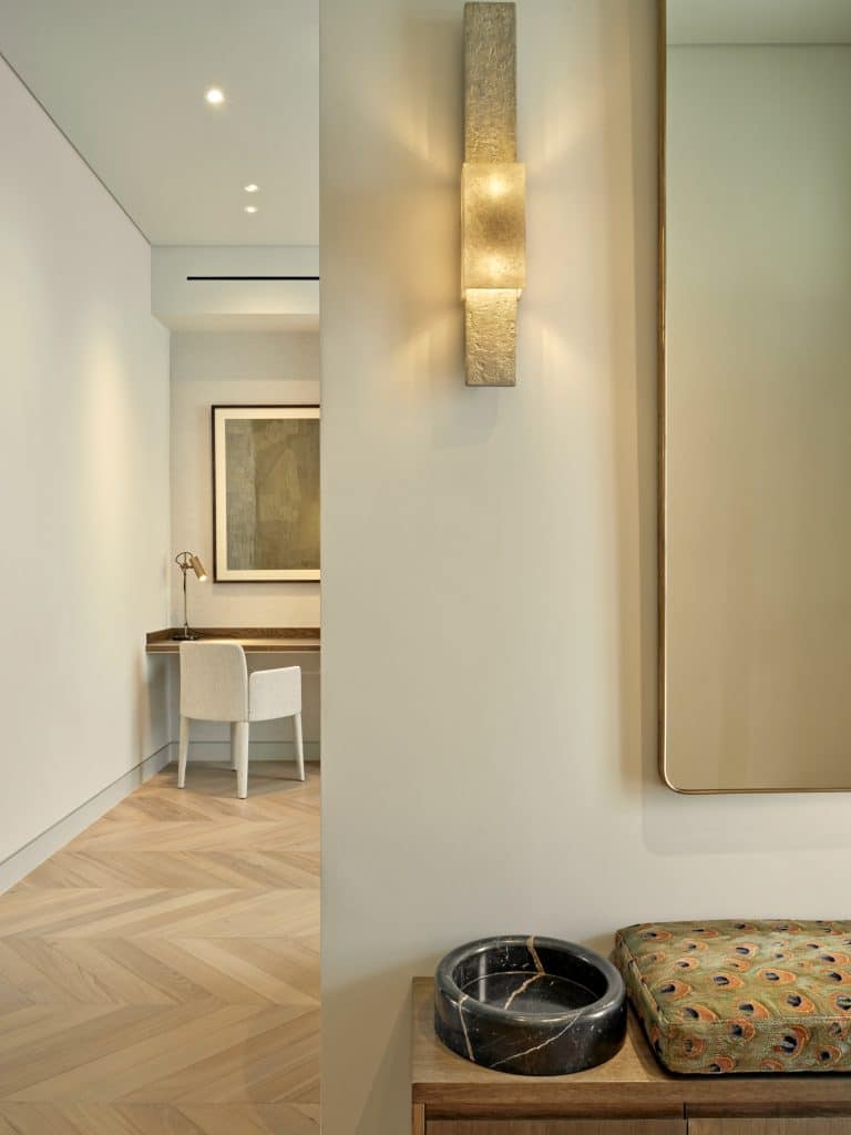 A zoomed-in image, unveiling an open plan space. The light wood fishbone flooring and Crisp white walls sets the stage , while a stylish wooden console takes centre stage, adorned with a luxurious pet water bowl made from black marble with delicate white veins. Nestled beside it, a pet bed cushion crafted out of brilliant peacock feather print fabric. Above the console, a grand rectangular mirror with gold rims reflects the beauty and style. Accompanying the mirror a unique structured stone light appointed on the wall, emitting a cosy warm glow. Behind the console, a glimpse into the study area reveals a captivating dark brown built-in desk, accompanied by a cream chair and captivating artwork adorning the walls.