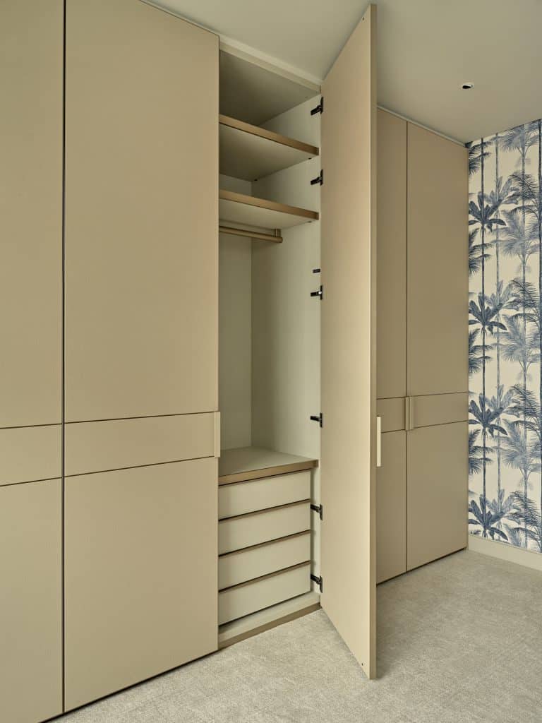 This luxurious bedroom/dressing room offers a harmonious blend of elegance and functionality. The wardrobe joinery is adorned in a soothing beige leather, with one open door revealing a well-organised selection of storage options. A plush beige woven carpet enhances the comfort and style of the space. The walls are adorned with a striking blue and white wallpaper featuring palm trees and plants, creating a captivating tropical ambiance. This combination of tasteful design elements transforms the room into a serene and sophisticated retreat.