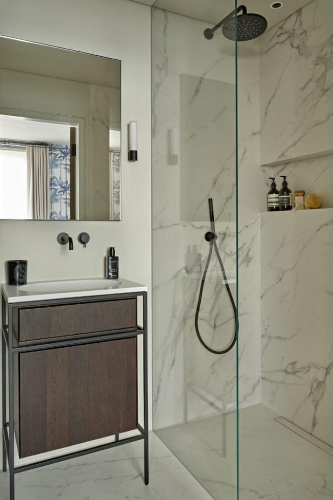 Luxury bathroom adorned with white marble featuring grey veins. The shower has a sleek glass screen and black hardware, complemented by a built-in shelf adorned with bathroom accessories. To the left, a white sink with a dark wood and black metal base, accompanied by a wall-mounted mirror and subtle wall lights on either side,