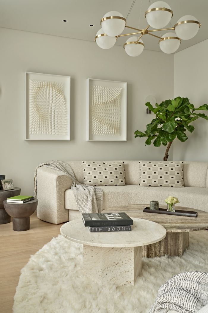 This luxurious living room features a corner space adorned with a curved white, textured sofa. Three matching white cushions and a white throw add comfort and elegance. A fluffy curved rug lies in front of the sofa, hosting two marble tables in different natural tones. Adjacent to the sofa, two black side tables display books and framed photos. Adorning the wall are two white textured art pieces, while behind the sofa stands a tall, vibrant green plant, adding a touch of nature to the space.