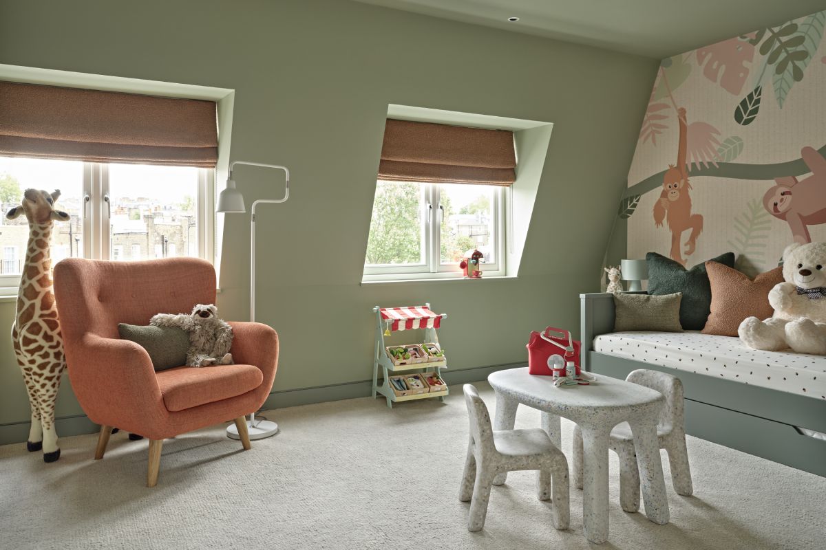 A luxury kids' playroom, transformed into a vibrant jungle paradise. A green trundle bed adorned with burnt orange and green cushions is accompanied by stuffed jungle animals. The walls are adorned with wallpaper showcasing lush jungle scenery and playful animals. The green-painted walls create a lively atmosphere. Burnt orange blinds adorn the window, adding a pop of colour. Green old suitcase-style side tables with matching lamps provide storage and illumination. A toy food shop and toy coffee machine offer endless opportunities for creative play. A children's drawing table with two seats inspires artistic exploration. This jungle-themed playroom is a haven of fun and creativity for children to explore and let their imaginations roam. Adjacent to this, a cosy orange armchair accompanied by a small green accent cushion and a toy sloth, with a white floor lamp for reading carefully positioned . Behind the armchair a large toy giraffe stands tall bringing this cosy corner to life.