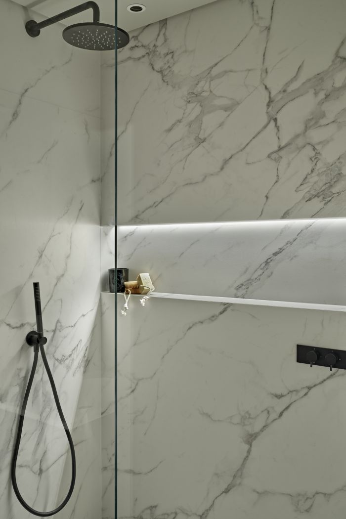 Guest bathroom with white marble effect porcelain tiles. the shower has a glass screen and black handwear, alongside a built in shelf accompanied with shower accessories.
