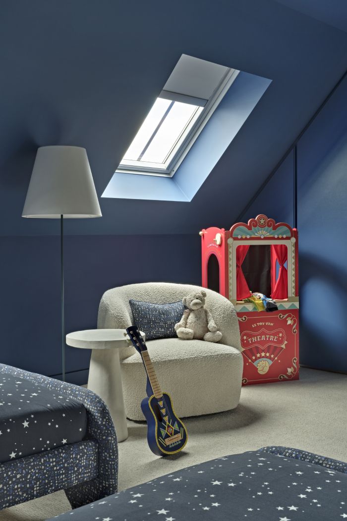 This luxurious kids' bedroom/playroom features calming blue painted walls illuminated by a skylight. A plush cream carpet adds comfort and warmth to the space. Two blue single beds adorned with bedding featuring white stars create a dreamy sleeping area. A cream fluffy, gwyneth boucle armchair with a matching blue cushion complements the fabric on the bed bases. A white tall side table holds a small blue guitar, while a white floor lamp provides additional lighting. In the corner of the room, a vibrant red mini puppet theatre awaits imaginative play and storytelling adventures.
