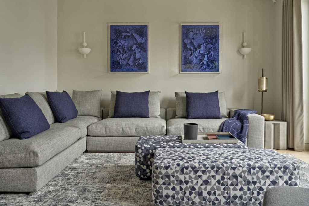 This luxurious living room where comfort meets elegance. The centerpiece is a large grey corner sofa adorned with plush cushions in various shades of grey and royal blue, providing both style and relaxation. A stunning blue/grey rug anchors the space, adding texture and depth. Two patterned ottomans, one featuring a convenient tray, offer versatility and extra seating. Enhancing the ambiance, two striking royal blue 3D framed art pieces take centre stage on the wall, beautifully complemented by unique light fixtures on either side
