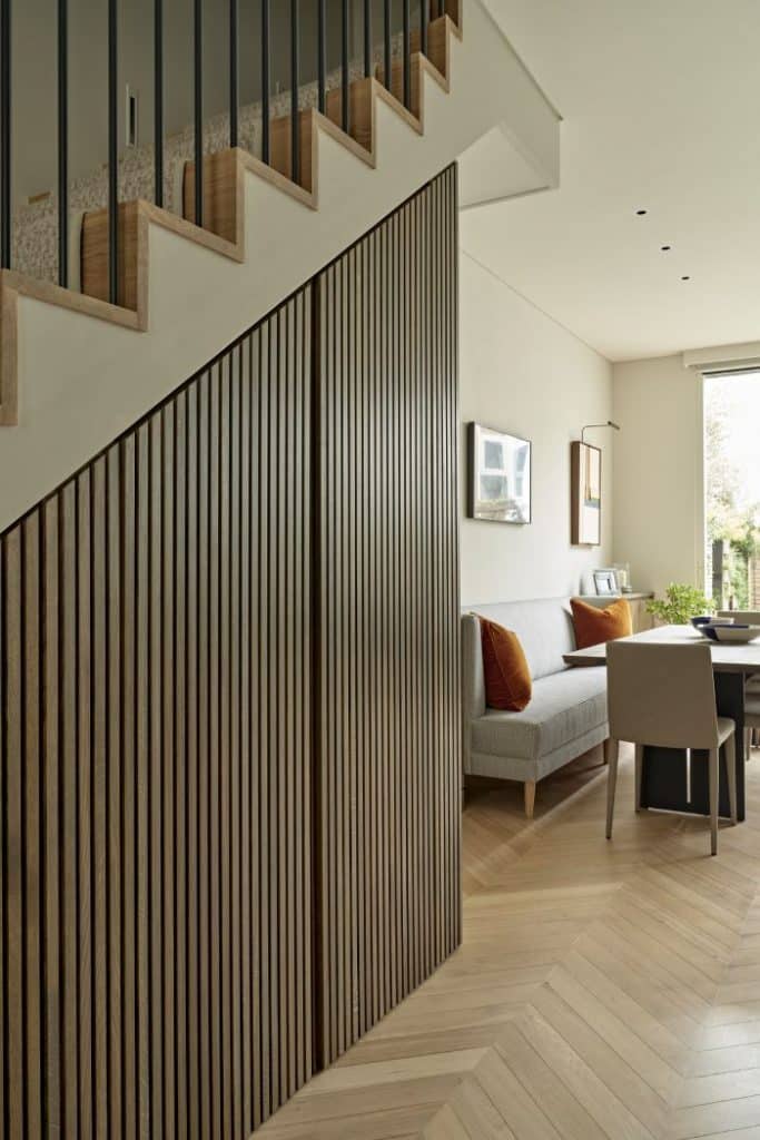 This zoomed-in image unravels the beauty of a luxury stairway. Thin wood panelling graces the wall beneath the stairs, concealing hidden storage that enhances both functionality and aesthetics. The stairs themselves are crafted from light wood and are adorned with beige carpet. Beyond the stairs lies an inviting dining space, featuring light wood herringbone flooring and pristine white walls and ceiling. A sophisticated grey dining couch with velvet orange cushions, while a light wood dining table adorned with tasteful bowls takes centre stage. A white chair stands at the end of the table. Above the couch, two captivating pieces of art captivate the senses, infusing the space with artistic allure.