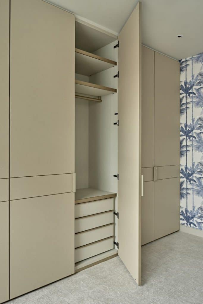 This luxurious bedroom/dressing room offers a harmonious blend of elegance and functionality. The wardrobe joinery is adorned in a soothing beige leather, with one open door revealing a well-organised selection of storage options. A plush beige woven carpet enhances the comfort and style of the space. The walls are adorned with a striking blue and white wallpaper featuring palm trees and plants, creating a captivating tropical ambiance. This combination of tasteful design elements transforms the room into a serene and sophisticated retreat.