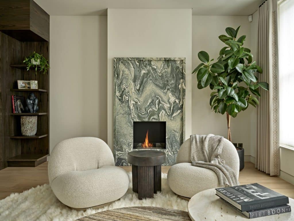 The luxury living room exudes elegance and sophistication in every detail. Positioned on a circular white fluffy rug, two high-end armchairs with textured white upholstery take center stage. A green veined marble fireplace stands proudly, adding a touch of grandeur. A stone side table complements the fireplace, offering a perfect spot for decorative items. In one corner, a tall green plant adds a fresh and natural element to the room. Dark wood corner shelves showcase an impressive collection of books and ornaments. Two marble coffee tables in varying natural tones provide both functionality and style, adorned with carefully placed books. The light wood floor adds warmth and enhances the room's airy atmosphere, while a window invites in natural light, completing this luxurious residential space.