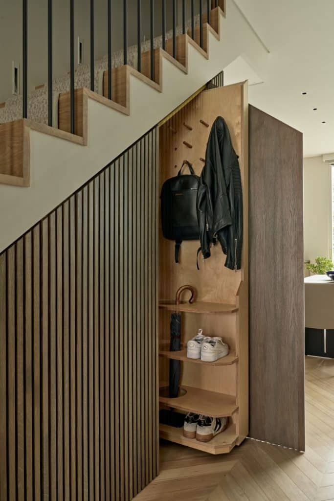 This luxurious under stairs showcases smart and efficient use of space. The slim paneled under stairs joinery features a pull-out storage unit, providing a practical solution for storing shoes, jackets, and bags. The light wood herringbone flooring harmonises with the light wood stairs, creating a cohesive and visually appealing look. A cream carpet running upstairs adds a touch of warmth and elegance. This cleverly designed under stairs room maximises functionality while maintaining a stylish and sophisticated atmosphere.