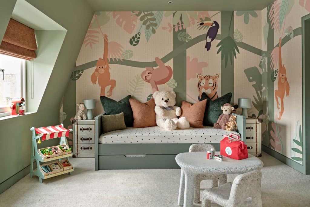 Welcome to the luxury kids' playroom, transformed into a vibrant jungle paradise. A green trundle bed adorned with burnt orange and green cushions is accompanied by stuffed jungle animals. The walls are adorned with wallpaper showcasing lush jungle scenery and playful animals. The green-painted walls create a lively atmosphere. Burnt orange blinds adorn the window, adding a pop of colour. Green old suitcase-style side tables with matching lamps provide storage and illumination. A toy food shop and toy coffee machine offer endless opportunities for creative play. A children's drawing table with two seats inspires artistic exploration. This jungle-themed playroom is a haven of fun and creativity for children to explore and let their imaginations roam.