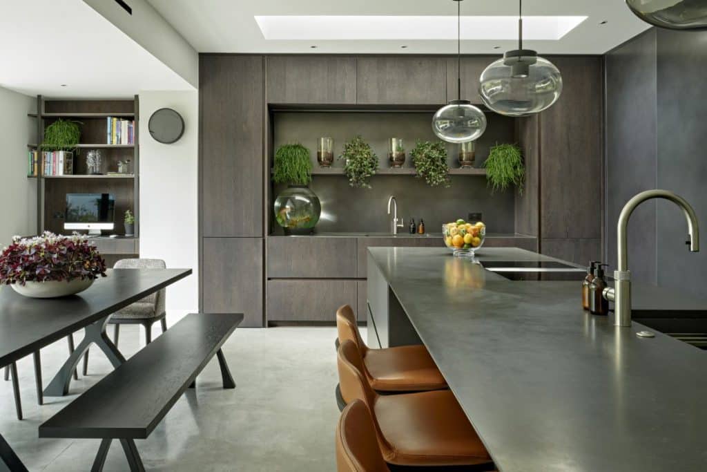 A luxury kitchen. The white ceiling adorned with skylights fills the space with natural light, illuminating the light grey stone floor. Along the back wall, dark wood joinery with kitchen cabinets and a built-in countertop with a sleek sink and a fish tank. Above the sink, shelves adorned with lush green plants. In the centre of the room, a kitchen island crafted from black slate tiles offers a functional workspace, with a built-in sink and hobs. Alongside the island, a dark wood dining table with benches, with a large potted plant adds a vibrant touch.