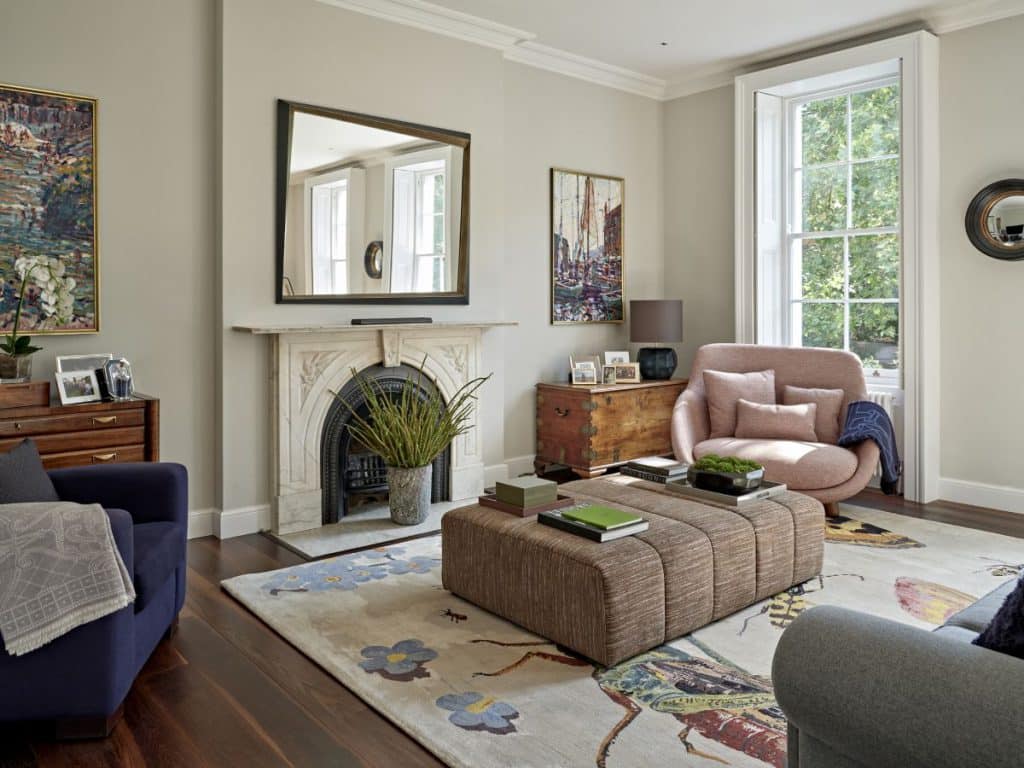 This luxury living room boasts a big window, allowing natural light to illuminate the space. A light burgundy love couch with matching cushions and a dark blue throw exudes comfort and style. In another corner, a dark blue armchair with a grey throw provides additional seating. The dark wood floor adds warmth and elegance to the room. A rug adorned with bugs and flowers becomes a centerpiece, featuring a brown textured ottoman with books. A grey sofa with dark blue cushions complements the layout. Across the room, a beautiful fireplace with a mantel, adorned with a plant, creates a cosy focal point. A mirror hangs above the fireplace, reflecting the room's beauty. Wooden corner tables on either side of the fireplace display lamps, framed pictures, and plants, with art gracefully hung above them.