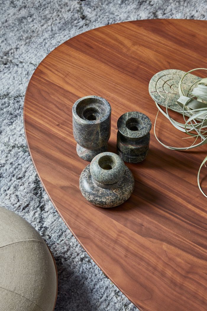 Up close image of a wooden toped coffee table creates a warm focal point in this living room with three cohesive stone candle holders on display, next to a wispy green plant which brings the scene to life. This coffee table is adorned on a grey texture, fluffy rug, alongside a clay green, wooden based pouffe .