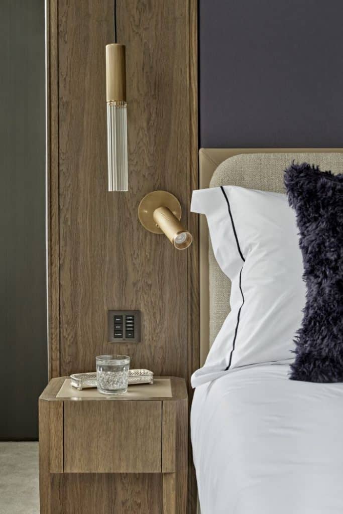 This high-end bedroom showcases a luxurious ambiance with its carefully curated design elements. A dark wood room divider serves as an elegant backdrop to the double bed, while matching bedside tables add a cohesive touch. The grey/blue fabric wall panelling behind the bed's headboard creates a soothing focal point. A natural cushion headboard enhances the comfort and style of the double bed, complemented by white cotton bedsheets with blue/grey detail edging. Fluffy blue/grey accent cushions add a touch of plushness, while hanging lights on either side of the bed provide atmospheric illumination alongside wall-mounted book lights.