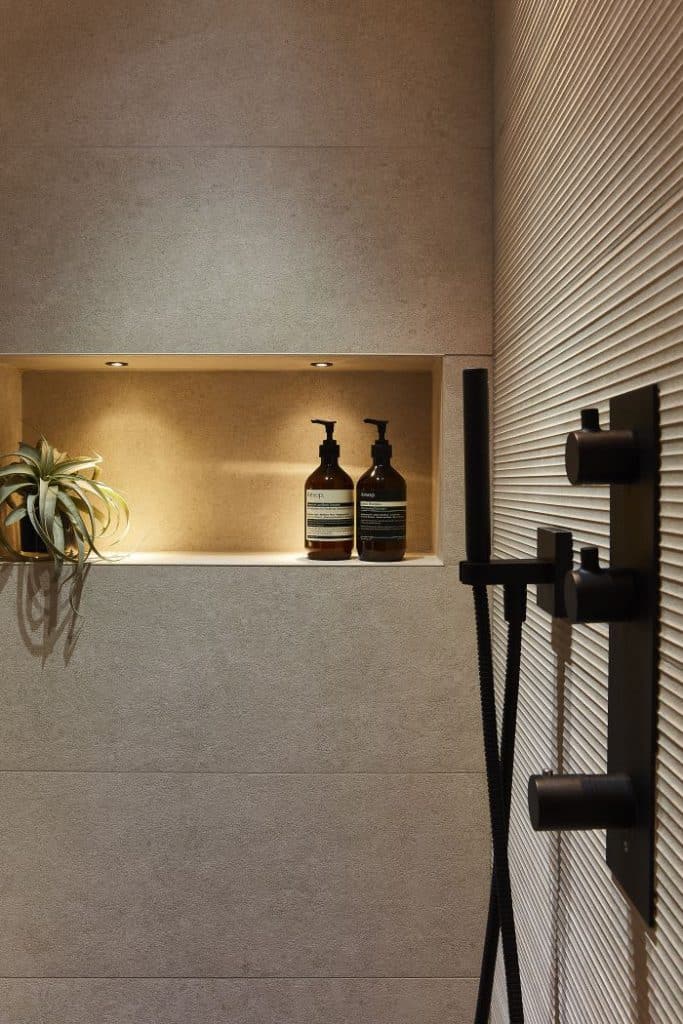 This luxurious bathroom features a sleek and contemporary design. The shower is adorned with grey stone walls and a built-in shelf, beautifully illuminated by John Cullen lighting. On the shelf, bottles of body wash are neatly displayed alongside a small potted plant, adding a touch of nature. The opposite wall showcases textured grey tiles, with a black shower head and panel creating a stylish contrast.