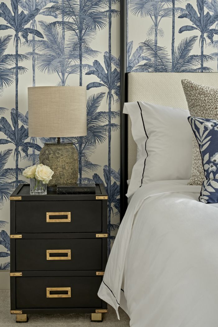 Old world-inspired luxury double bedroom exudes sophistication and elegance. A steel four-poster bed frame serves as a striking centrepiece, complemented by a cream headboard and crisp white bed sheets. Cream patterned accent cushions and blue accent cushions. The walls are adorned with blue and white wallpaper featuring palm trees, creating a serene tropical ambiance. Dark blue and gold bedside tables, adorned with white lamps inspired by Amelia Carter Interiors, add a luxurious touch.