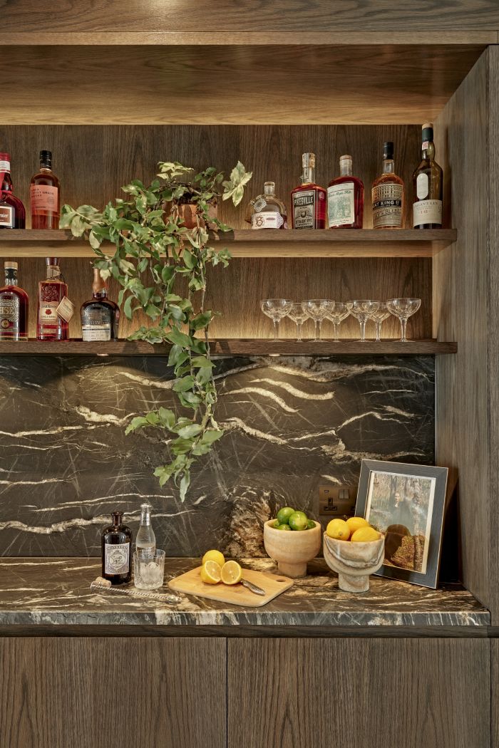 A luxurious modern home bar, designed for cocktail enthusiasts. The dark wood joinery features illuminated shelving, showcasing an array of meticulously displayed liquor bottles and glasses. A stunning grey marble countertop with elegant white veins adds a touch of sophistication. On the countertop, a selection of fresh lemons and limes are accompanied by a cutting board, ready for the creation of refreshing drinks. This luxury home bar seamlessly combines functionality and style, providing a sophisticated space for mixing and enjoying cocktails.