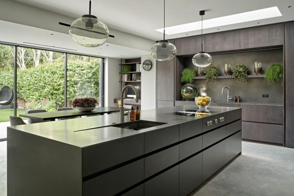 This luxury kitchen boasts a refined and modern aesthetic. Light grey stone-look flooring sets the foundation for the space. Dark wood kitchen cabinets and joinery, paired with a sleek slate black countertop and sink, create a striking contrast. Shelves adorned with vibrant plants above the sink add a touch of freshness. On the opposite wall, slate black cabinets A slate black island with hobs becomes a centrepiece, complemented by a large glass bowl filled with vibrant oranges, lemons, and limes. Above the island, large glass bulb lights illuminate the workspace. Expansive glass sliding doors and a skylight flood the kitchen with natural light, enhancing its luxurious ambiance.