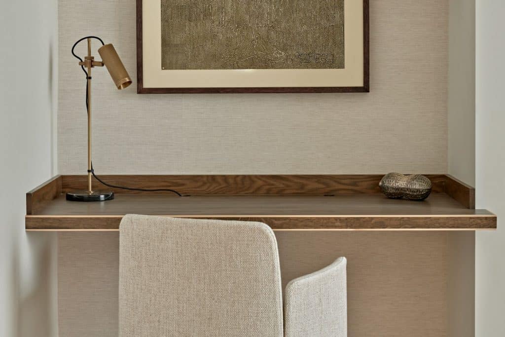 A stunning up-close section of the room showcases a dark wood desk seamlessly integrated into the wall. A beige textured fabric desk chair adds elegance and comfort. Next to the pristine white walls, a beige fabric panel wallpaper exudes sophistication. A gold spotlight desk lamp illuminates the workspace . Above the desk, a textured art piece is framed with a dark wood frame, creating a focal point of artistic beauty.