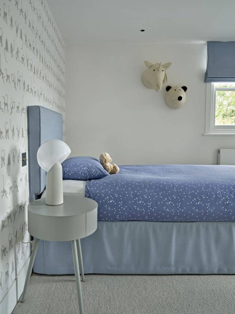 A luxury childrens bedroom, a blue based bed with matching headboard, complemented by delicate blue starred bed sheets accompanied by a small fluffy teddy bear adding a calm effect. To the side of the bed a sleek round side table with a pull out drawer and a quirky white lamp. On the wall are whimsical stuffed animals heads hanging on children's bedroom wall, a fun cream unicorn and a matching bear head. in the background on another wall a monkey wallpaper, matching the animal theme.