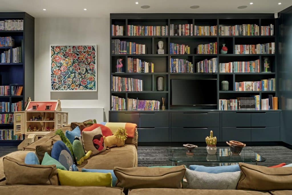 A luxurious and fun living room,where elegance meets playfulness. On one side, sleek dark grey joinery, with shelves and slide out drawers displaying an array of books and ornaments. A television stands alongside on the joinery. Adjacent to the joinery, a vibrant and colourful painting of flowers adds a cheerful touch, while underneath, a whimsical children's dollhouse. In the foreground, a cosy brown corner sofa, adorned with an assortment of playful animal-shaped cushions. A sleek glass coffee table takes centre stage, with a delightful plant and ornaments.