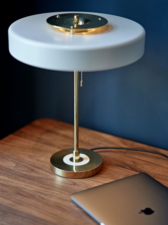 A zoomed in image of a work space located in a grand double bedroom. The image features a rich dark walnut wood desk with an elegant brass berk frank revolve table lamp crafted with a gold base, the lampshade in a cylindrical shape in white creates a graceful focal point, sitting next to the lamp, a MacBook, providing the space with functionality .