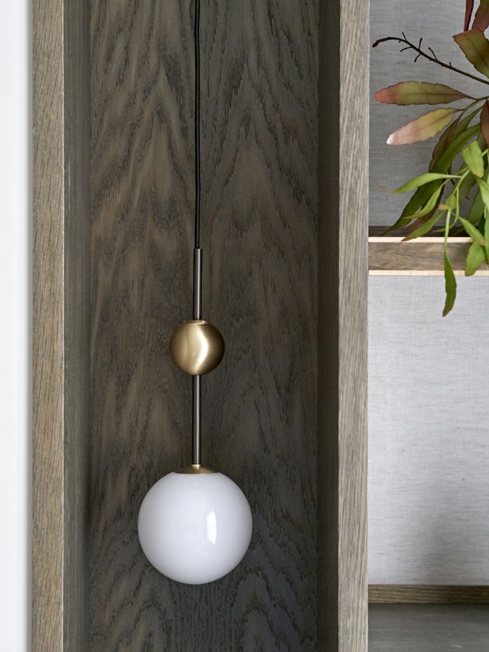 A zoomed-in image showcasing a captivating hanging light. Two elegant spheres adorn this pendant fixture, one as a light function, while the other serves as a gold decoration. Suspended within a dark wood joinery, this lighting piece creates a captivating focal point. Surrounding the light, carefully crafted shelves adorned with lush green plants add a touch of natural beauty.
