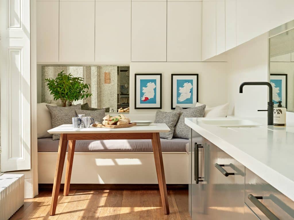 A cosy kitchen lounge area with an inviting atmosphere. The light wood floor sets a warm and tone. On the right, a white kitchen countertop with a sleek grey base becomes the focal point, accompanied by stylish cabinets on the walls above. Further into the space, a cosy lounge area, adorned with soft white and grey cushions. A wooden table with a clean white top serves as a relaxing place. Behind the lounge area, framed map images and a mirror add a touch of sophistication, with a lush green plant.
