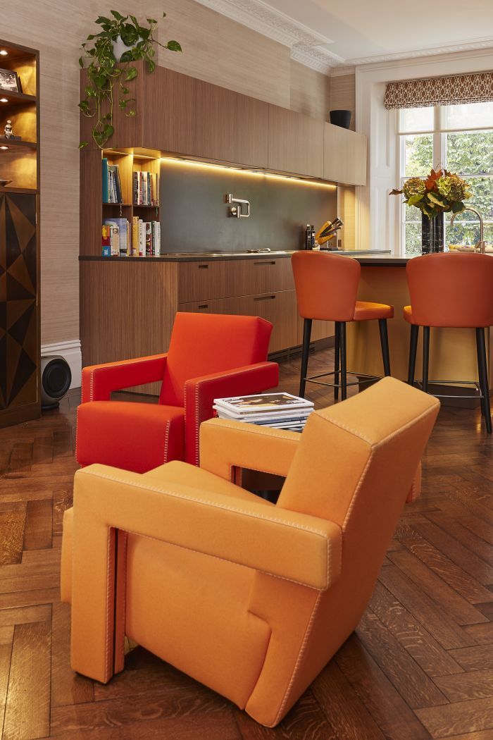 A luxurious open plan living room and kitchen.The herringbone wooden flooring sets the stage for a pair of Cassina Utrecht armchairs in bold red and orange , creating a vibrant focal point. As you glance beyond, you'll find a beautifully designed kitchen with light wood joinery and sleek grey stone countertops. Built-in cookbook shelves and a convenient pot filler add practicality to the space. At the heart of it all, a circular island with a yellow base and grey stone top, adorned with a vase of fresh flowers. Surrounding the island, orange chairs with black legs provide comfortable seating. Finally, a large window at the back floods the room in natural light.