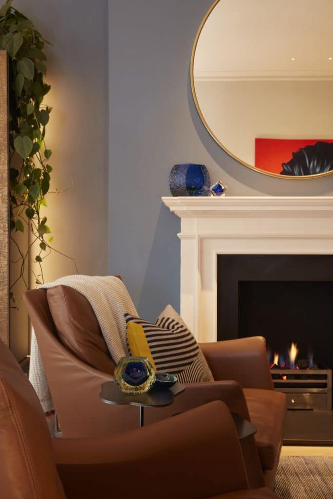 chesney limestone fireplace with a large circular mirror above it. In front of the fireplace a set of brown leather armchairs are seated.
