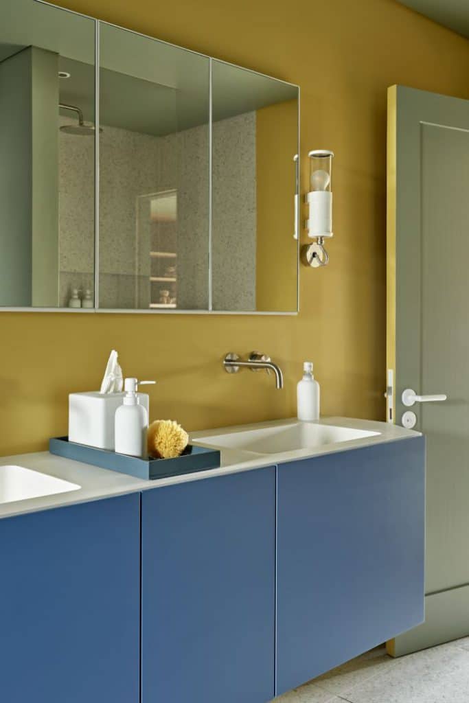 A captivating kids' bathroom. The vibrant colours instantly grab your attention, the yellow wall that sets the tone for a cheerful atmosphere. A sleek mirror accompanied by an industrial-style light fixture adds a touch of sophistication. Just below, a twin sink with a blue base takes centre stage, adorned with brass taps. The bathroom accessories accentuate the space, bringing in even more pops of colour.