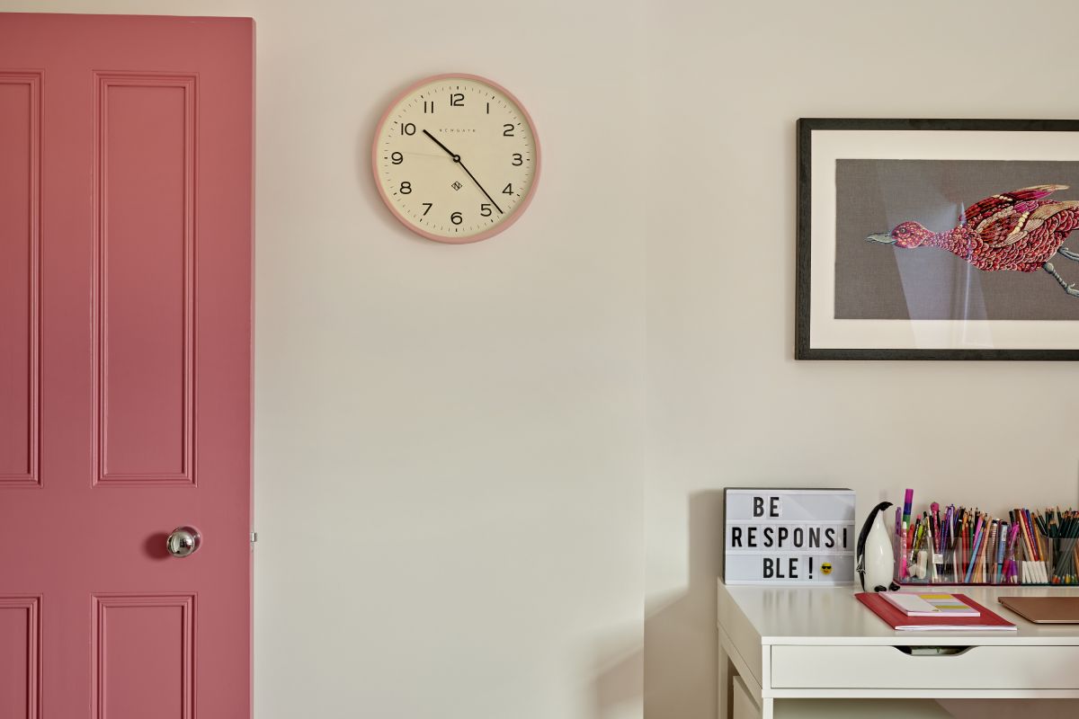 A zoomed in image of a luxurious pink girl's bedroom. The pink door welcomes you into the room. The walls, painted in pristine white, hangs a delicate pink clock. A white desk takes centre stage, adorned with a delightful collection of pens and books. Above the desk, a framed image of a pink bird, housed in a sleek black frame, captures attention with its graceful beauty.