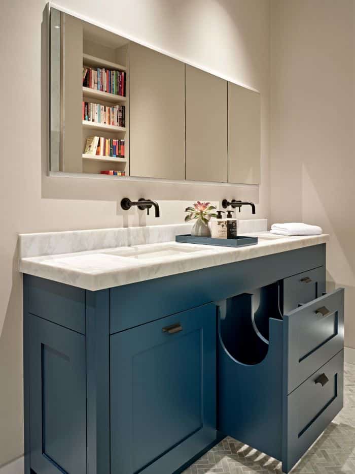 A luxurious bathroom that exudes elegance and functionality. The centrepiece of the space is a his and hers sink, with a pristine white marble countertop resting upon a beautifully crafted blue painted base. The sink is accompanied by pull-out drawers with lots of storage, ensuring a clutter-free environment. Black taps and stylish bathroom accessories add a touch of sophistication. Above the sinks, four panelled mirrors create a captivating display, reflecting the opposing wall adorned with shelves filled with a vibrant collection of colourful books.