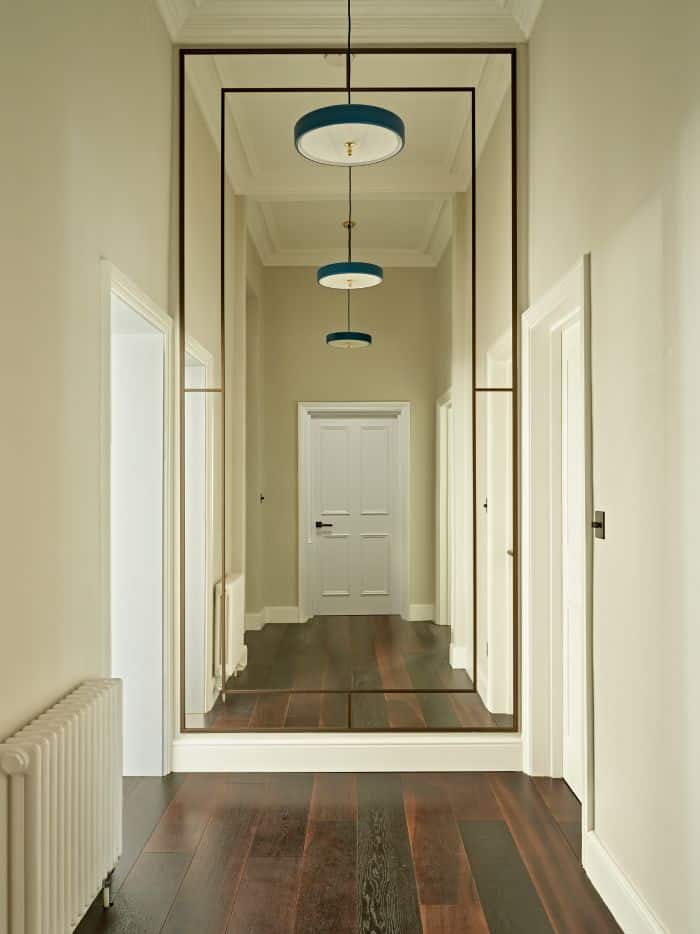 A luxurious hallway adorned with tall ceilings, cream walls, and exquisite dark wood flooring. The focal point of the image is a grand mirror with bronze details that stretches from floor to ceiling, capturing and reflecting the beauty of the space. The mirror reflects the door in the hallway and the stunning blue cylinder lights that hang from above. With an abundance of natural light streaming through, the hallway creates an elegant and welcoming ambiance.