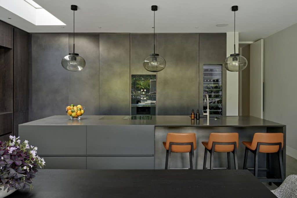 A luxurious Modulnova kitchen from Design Space London, where elegance and functionality meet. At the back, the dark grey marble kitchen joinery stands as a statement piece, featuring a built-in oven and a wine fridge. The centrepiece of the room is a kitchen island, matching the joinery's design. On one side, three brown leather stools with dark wood legs and a sink, while on the other side, cooking hobs and a vibrant display of citrus fruits in a glass bowl. Hanging over it, three glass globe lights. In the foreground, a dark wood dining table adorned with fresh flowers.