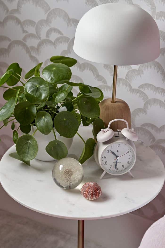 Zoomed-in image, captured from a child's bedroom. A white marble bedside table, adorned with a sleek white lamp with a wooden base. The table holds various personal accessories, including an alarm clock and a lush green plant, adding a touch of personality. Behind the table, the walls come alive with charming white cloud patterns on the wallpaper, creating a serene atmosphere.