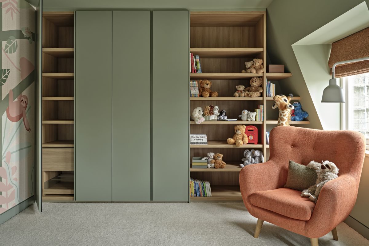 A luxurious children's playroom where creativity and comfort come together. The cream carpet sets the foundation for a cosy atmosphere, while the green walls add a touch of vibrancy. Along the back wall, bespoke joinery provides wardrobe space, alongside shelves that display a collection of colourful books and stuffed toys. A window adorned with textured orange blinds invites in natural light. In front of the window, a plush orange armchair with a green accent cushion becomes the perfect spot for reading, accompanied by a charming toy sloth. Standing tall behind the armchair, a playful toy giraffe adds a whimsical asset.