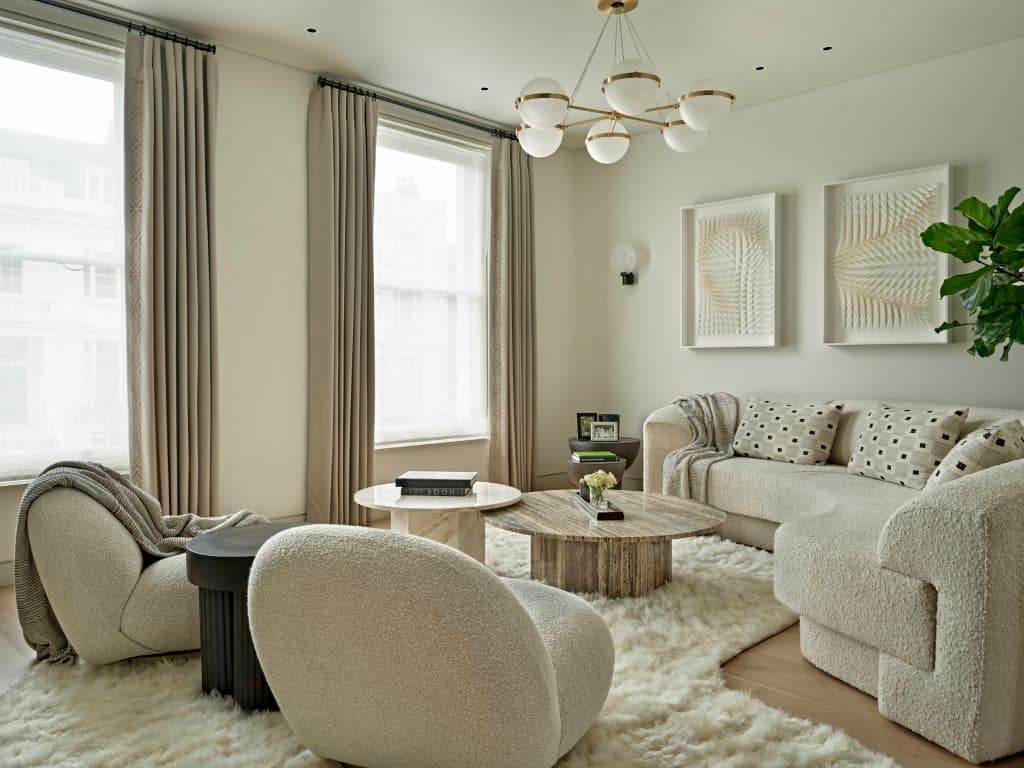 A luxury residential living room, where elegance meets comfort. Positioned on a curved white fluffy rug are two white, textured high-end armchairs, inviting you to relax in style. A corner sofa, matching the fabric of the armchairs, features curved edges for a touch of sophistication. Three simple patterned cushions adorn the sofa, adding a pop of color and coziness. A matching blanket drapes gracefully on the corner of the sofa. Two white textured art pieces hang on the white wall, adding visual interest and depth. A tall green plant in the corner brings a touch of nature indoors. Two marble coffee tables in different natural tones proudly display books on top. The light wood floor adds warmth and elegance to the room. Two windows, adorned with natural curtains, invite in abundant natural light, completing this luxurious living room.