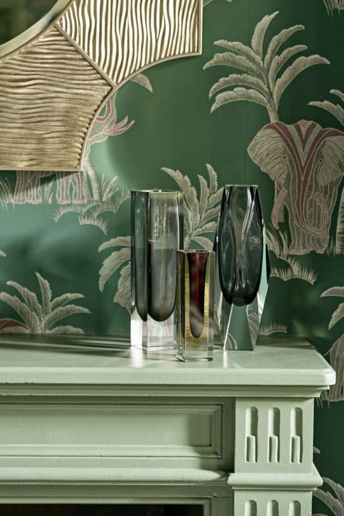 Up close image of three glamorous vases adorned on a green mantel piece, backing onto delicate green wallpaper showcasing images of palm trees and elephants. Hanging above is an extravagant gold mirror.