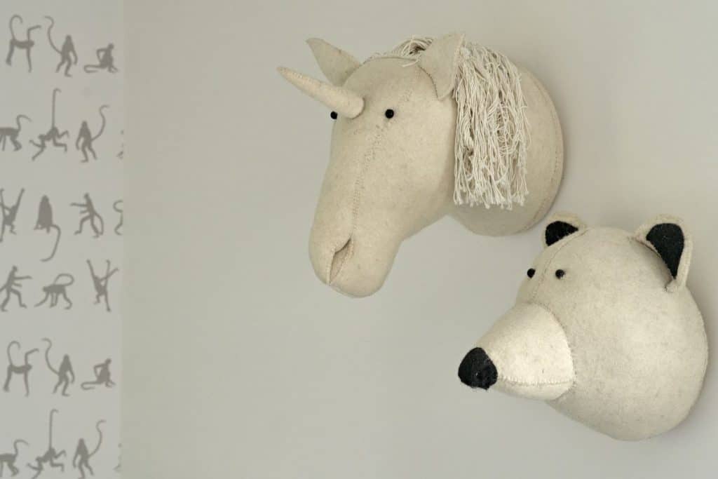 Close up image of whimsical stuffed animals heads hanging on children's bedroom wall, a fun cream unicorn and a matching bear head. in the background on another wall a monkey wallpaper, matching the animal theme.
