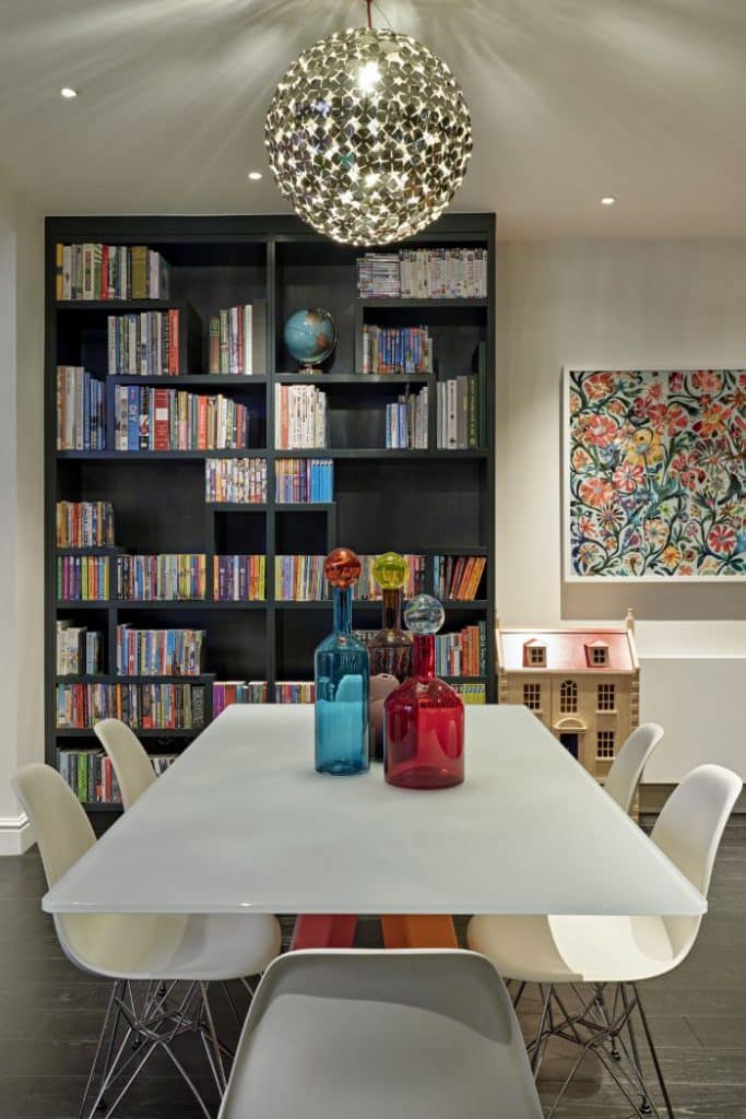 A luxurious dining room. The centrepiece is of a white glass tables surrounded by six sleek white chairs, creating a chic atmosphere. Adorning the tables are three stunning glass vases in different vibrant colours.. Behind the dining area, a large dark grey bookcase showcases an extensive collection of beautifully arranged multi-colored books.To the right of the bookcase, a colourful painting of flowers in a white frame, complemented by a charming children's dolls house below.