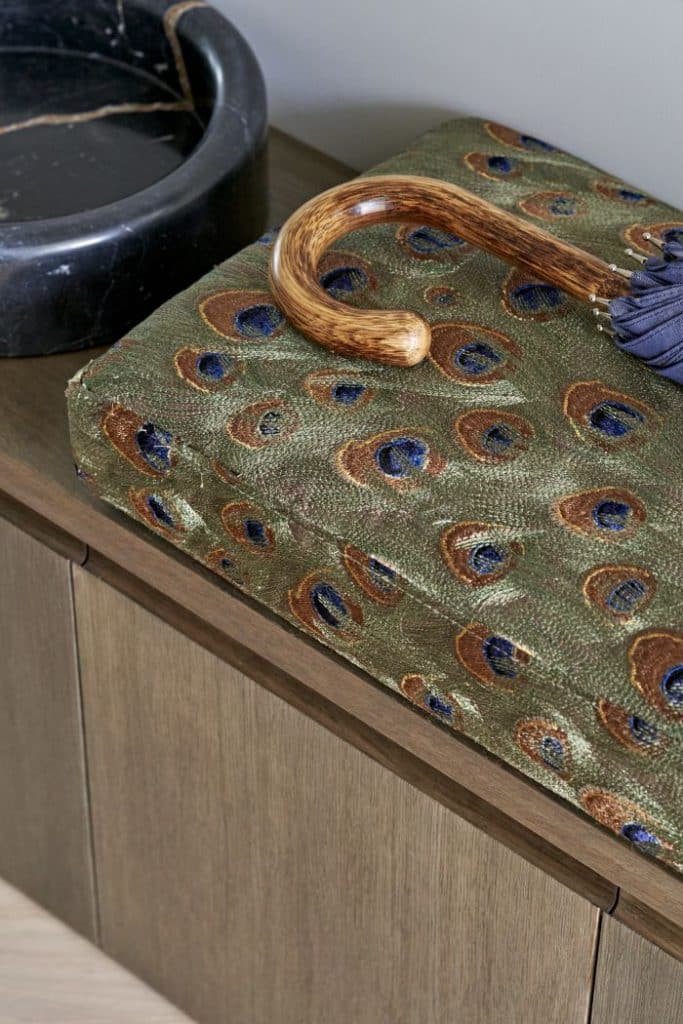 An up-close image of a luxurious area. A dark wood bench with drawers exudes functionality. Adorned with a plush cushion made out of a stunning peacock feather pattern. Alongside the cushion rests a wooden-handled umbrella. Adding a touch of sophistication, a black marble pet bowl sits nearby.
