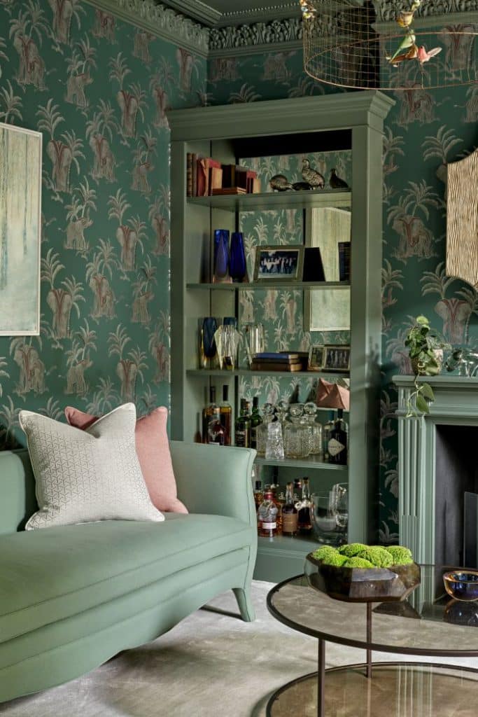 A luxurious green living room. The light green carpet and walls adorned with palm trees and elephants wallpaper create a soothing ambiance. A fireplace stands on one wall, complemented by a light green mantelpiece and a golden mirror above. On either side of the fireplace, sets of green shelves with mirrored backs display treasured books, picture frames, and personal accessories. A plush green couch with cream and pink cushions invites relaxation, while a framed painting hangs above.