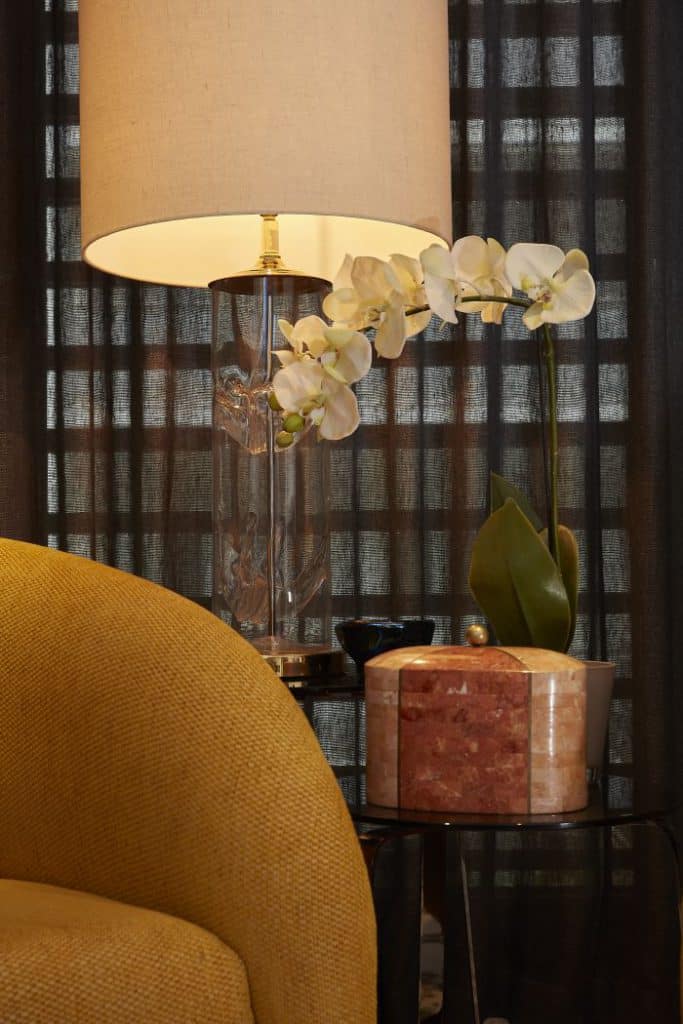 A close up shot showing the profile of a yellow upholstered chair, antique lamp and side table in the principle bedroom.
