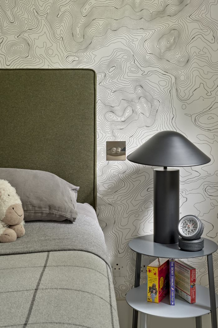 A luxurious children's bedroom, captured in a close up image. The walls are draped with a captivating black and white geographical map wallpaper, adorned with a silver light switch. A deep green headboard serves as the focal point. The bed is dressed in cosy grey sheets, accompanied by a cuddly stuffed toy resting on top. A sleek grey bedside table showcases a black bedside lamp with a whimsical mushroom-shaped lampshade. Books and accessories complete the table.