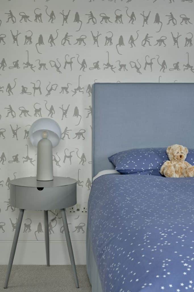 A luxury childrens bedroom, a blue based bed with matching headboard, complemented by delicate blue starred bed sheets accompanied by a small fluffy teddy bear adding a calm effect. To the side of the bed a sleek round side table with a pull out drawer and a quirky white lamp. The walls behind, the bed are draped in monkey themed wallpaper.