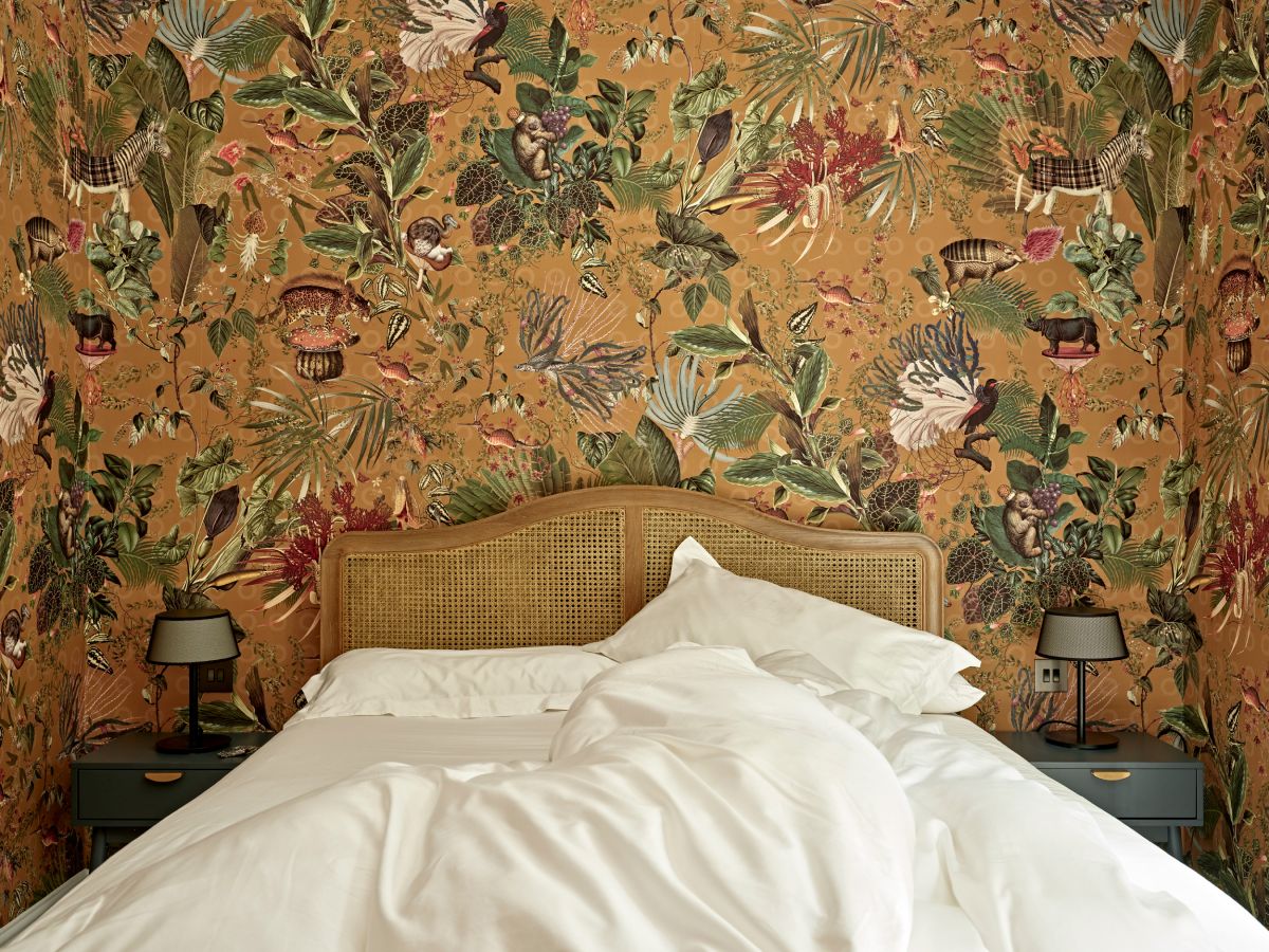 Close up image of a corner of a vibrant bedroom. The wall captures attention with its unique wallpaper from 'Arte International,' adorned with a captivating display of animals, plants, and flowers. Complementing the wallpaper, the wooden bed base with rattan headboard, accompanied by fresh white bed sheets create an abundance of calm. Nestled either side of the bed, blue elegant side tables with brass handles and sliding drawers add functionality. Adorned upon the side tables are vintage lamps emitting a soft warm glow bringing cosyness to the space.