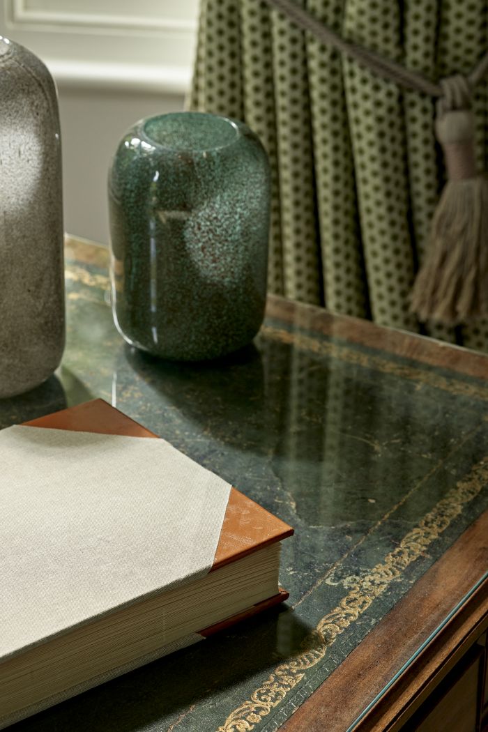The beauty of this luxury table. The close-up image shows a mesmerising combination of green marble, with intricate gold detailing and framed with dark wood. Positioned on the table, two elegant green vases, complemented by a fabric-covered book. Behind this setting, a set of green spotted curtains.